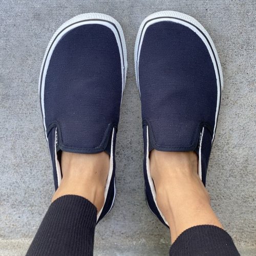 A photo of Bohempia Velik slip on sneakers made from canvas and rubber soles. The sneakers are a black color and white soles with a black stripe around the sole. A woman is shown above from the mid leg down wearing black joggers and the velik sneakers standing concrete steps. #color_black-white