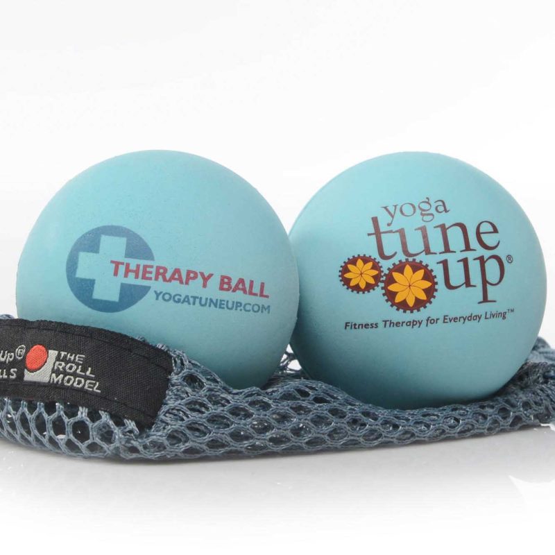 A photo of Tune Up therapy balls made from rubber. The therapy balls are a light blue color with therapy ball written on one side and yoga tune up on the other. Both balls are shown sitting on a gray mesh bag against a white background.