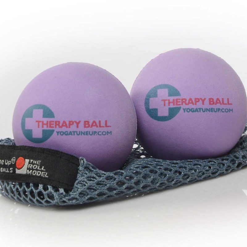 A photo of Tune Up therapy balls made from rubber. The therapy balls are a light purple color with therapy ball written on one side and yoga tune up on the other. Both balls are shown sitting on a gray mesh bag against a white background.