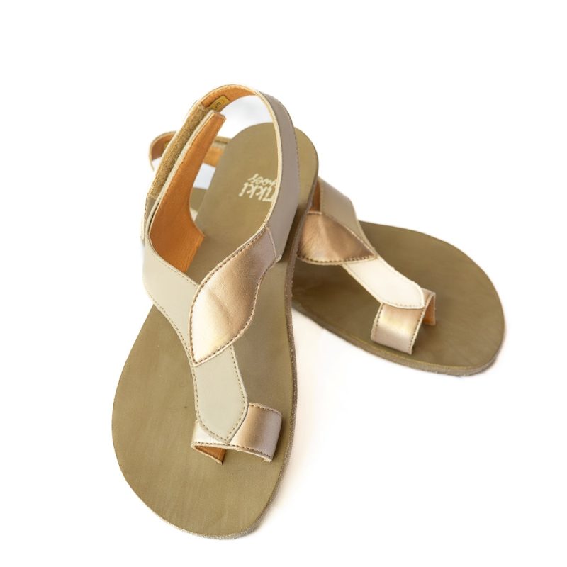 A photo of tan Tikki Soul leather sandals with a tan footbed and cream sole. Three leaf-shaped straps connect starting at a light gold toe ring, followed by a light tan piece, then another light gold piece surrounding the back of the foot and coneecting to a velcro closure at the outside of the ankle. Left shoe is shoen here from the front propped up on the right shoe which is facing right against a white background. #color_shell-vegan